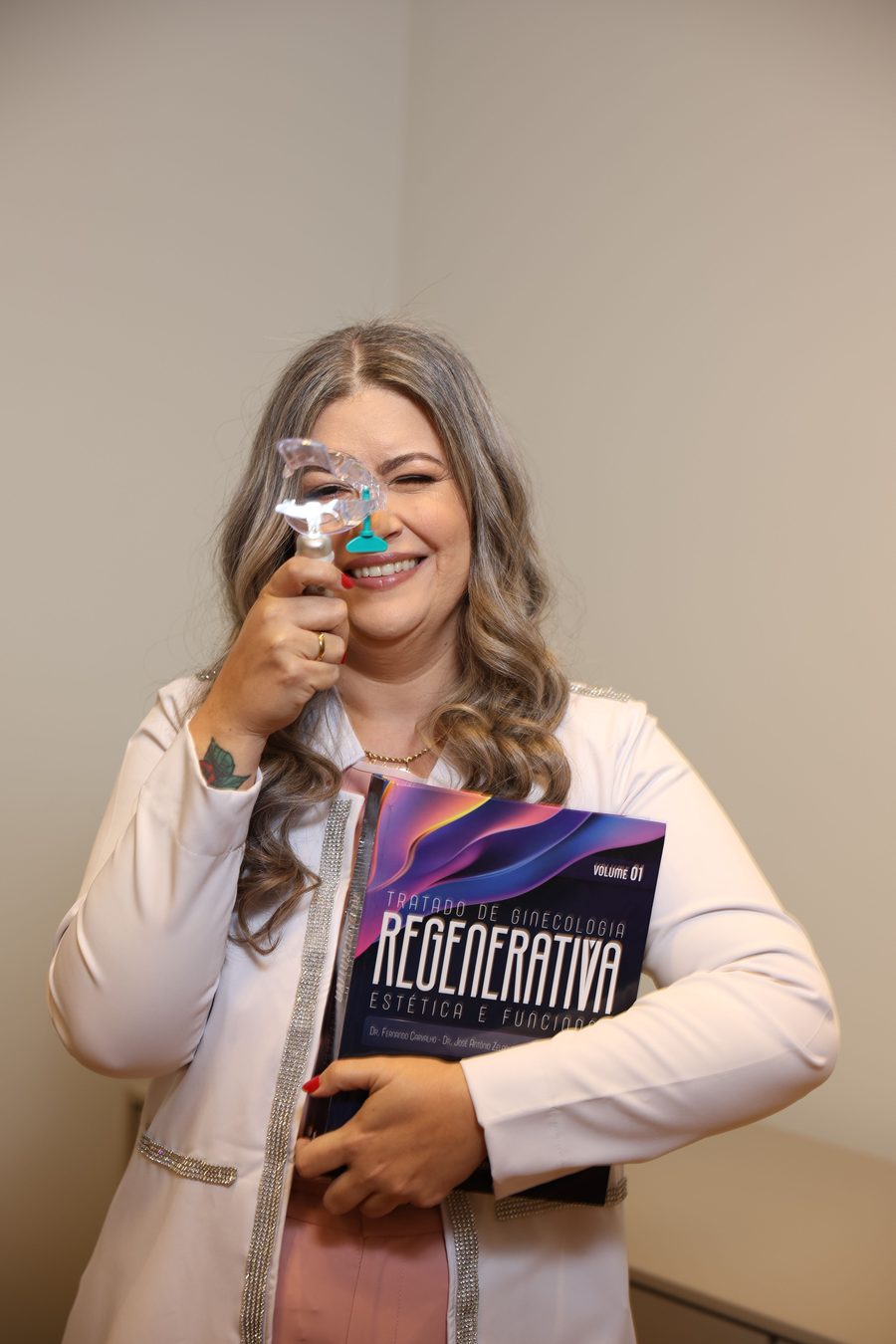 Dra. Julienni Arvelos sorrindo, de jaleco, segurando o Tratado de Ginecologia Regenerativa